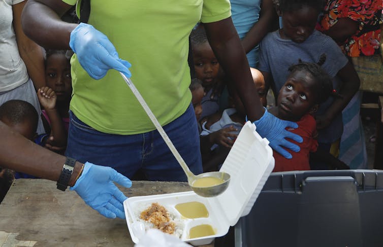 A waiter ladles soup into a container as children line up to receive food at a shelter.