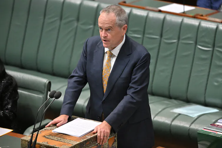 man stands at lectern in parliament interior with green leather seats