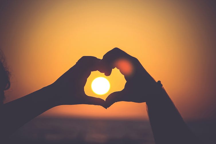 Two hands make a heart shape in front of a sunset.