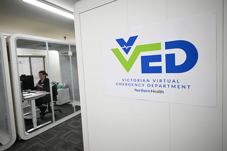Sign reads: VED Virtual Emergency Department. Woman sits nearby at desk in windowed cubicle