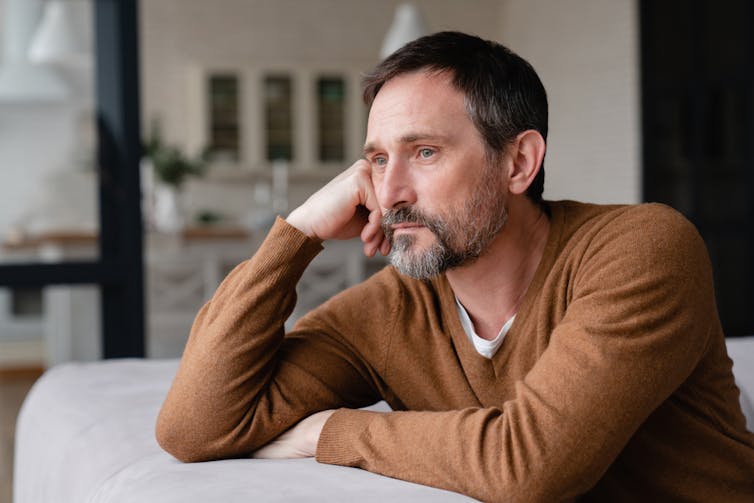 Middle-aged man looking sad, leaning on sofa, staring into distance.