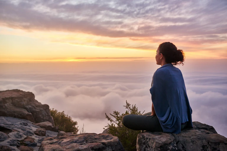 Photo of a woman sitting on top of a mountain looking out a sunrise over clouds.