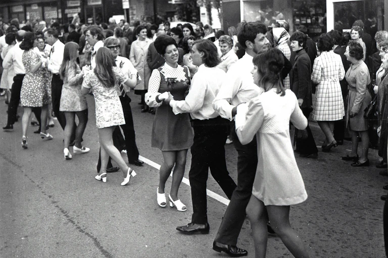 An archival black and white photograph of young people dancing in the street.