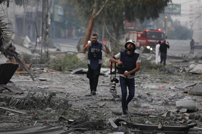 Two journalists walking through an area where buildings have been destroyed in Gaza.