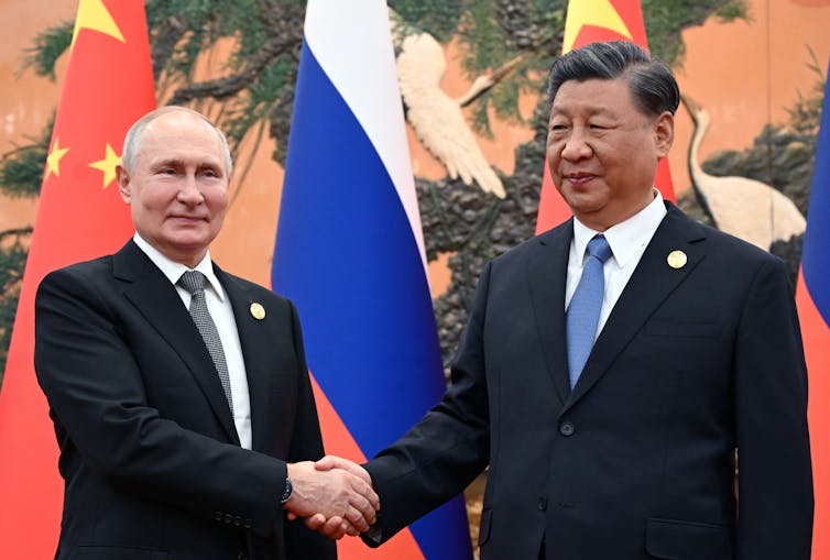Putin and Xi shaking hands in front of a wall draped in Russian and Chinese flags.
