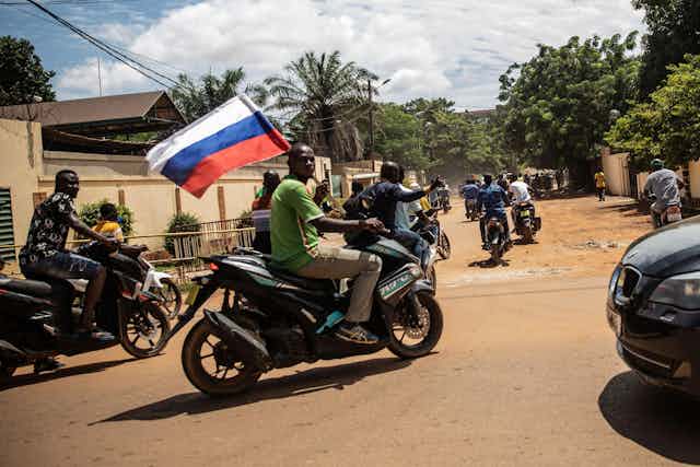 A group of men on motorbikes driving through a dusty street holding a Russian flag.