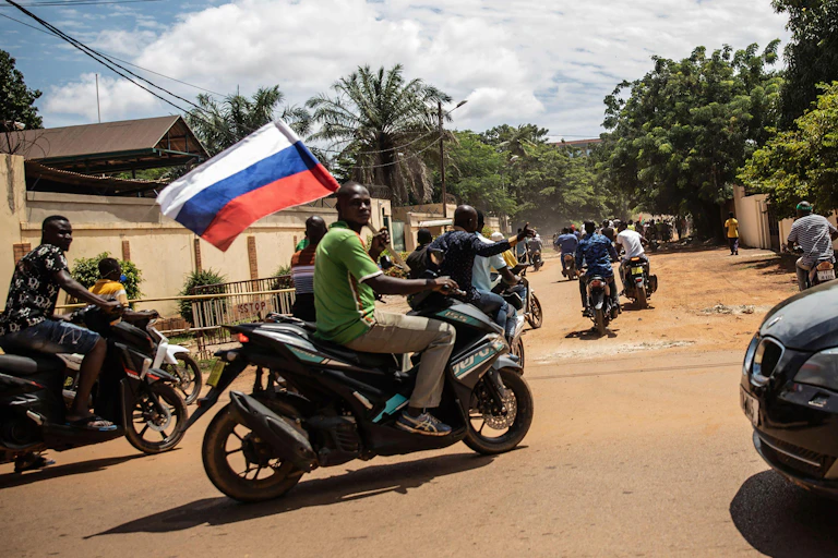 A group of men on motorbikes driving through a dusty street holding a Russian flag.