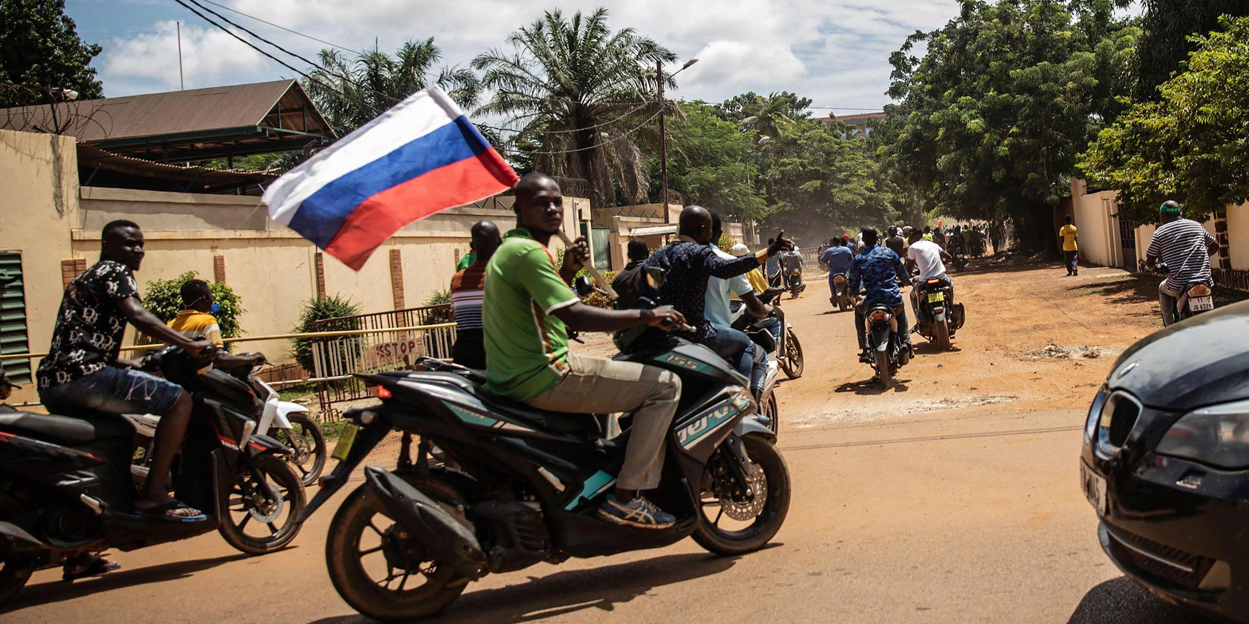 A group of men on motorbikes driving through a dusty street holding a Russian flag.