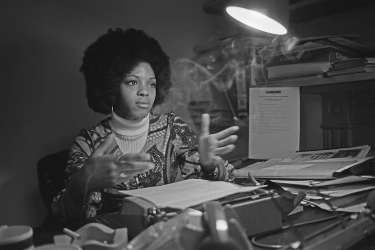 A young woman with an Afro sits at a desk piled with manuscripts, gesturing with her hands.