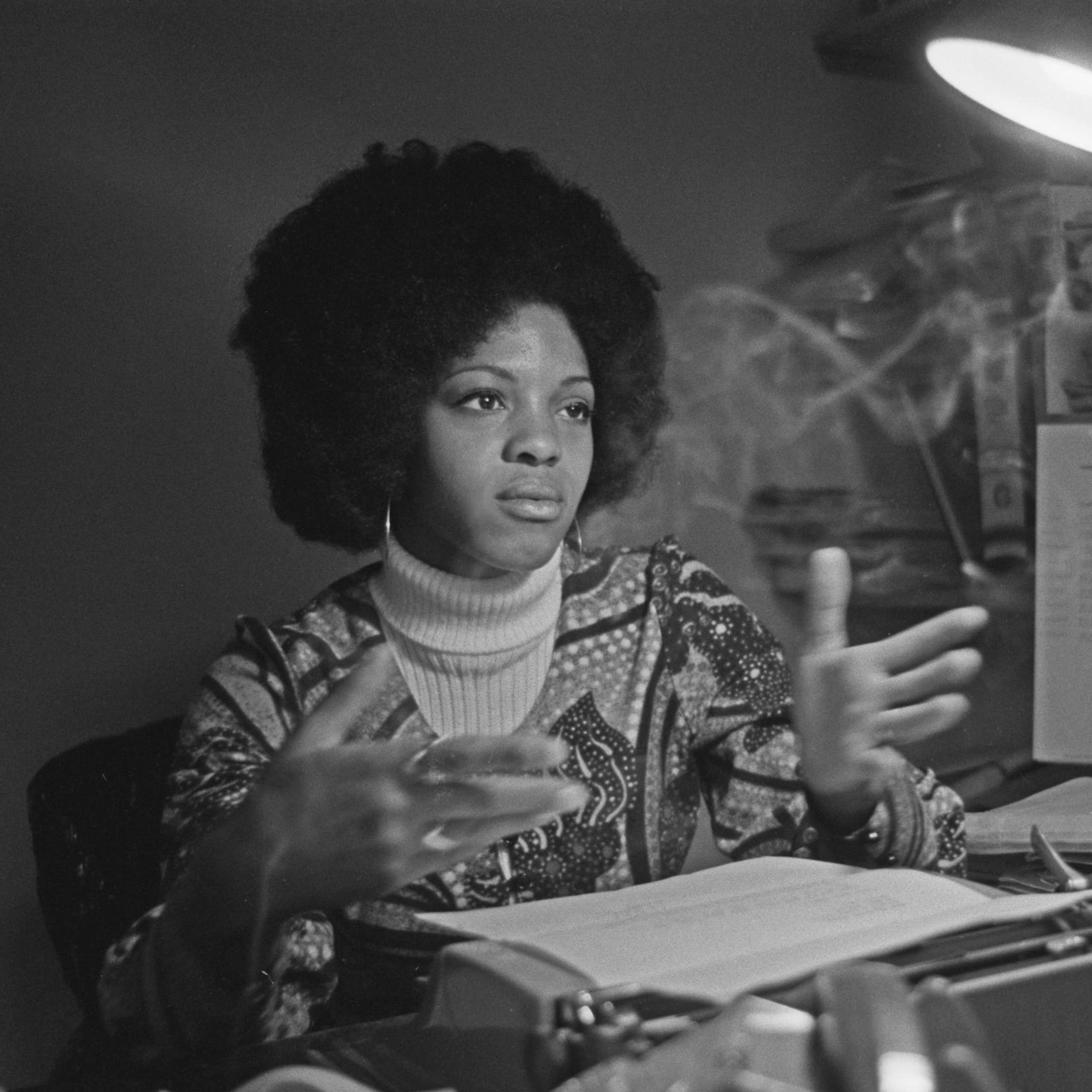 A young woman with an Afro sits at a desk piled with manuscripts, gesturing with her hands.