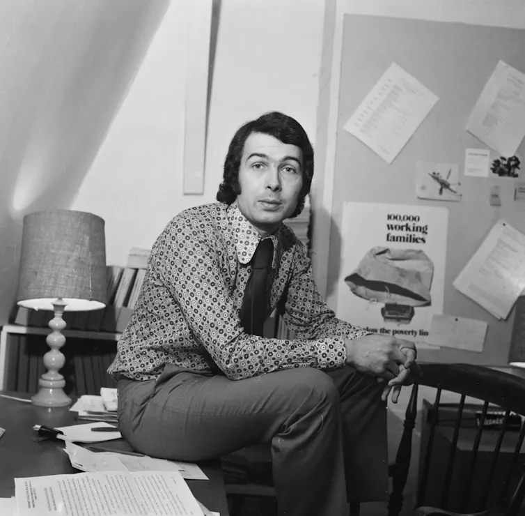 A black and white photo of a young Frank Field, sitting on a desk with his feet on a chair. Behind him is a poster that reads '100,000 working families below the poverty line'.