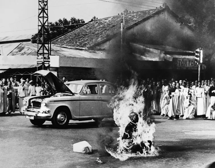 A black and white image of a Buddhist monk setting himself on fire on a road with a crowd watching on.