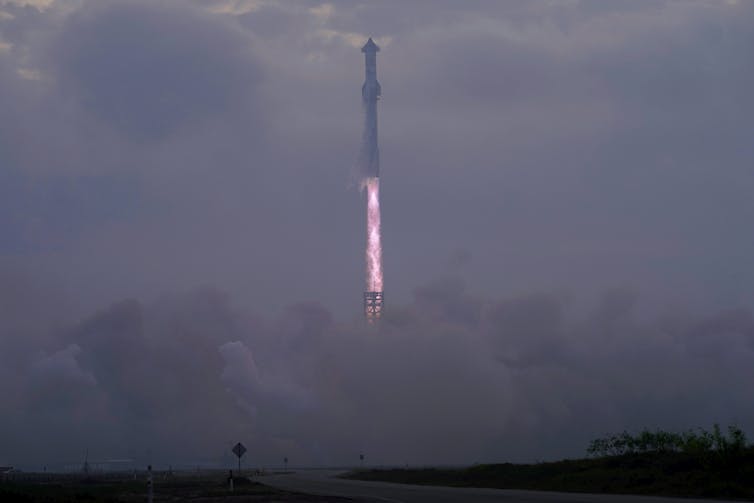 A long, cylindrical rocket with a plume of flames rising from the end shoots into the cloudy sky.