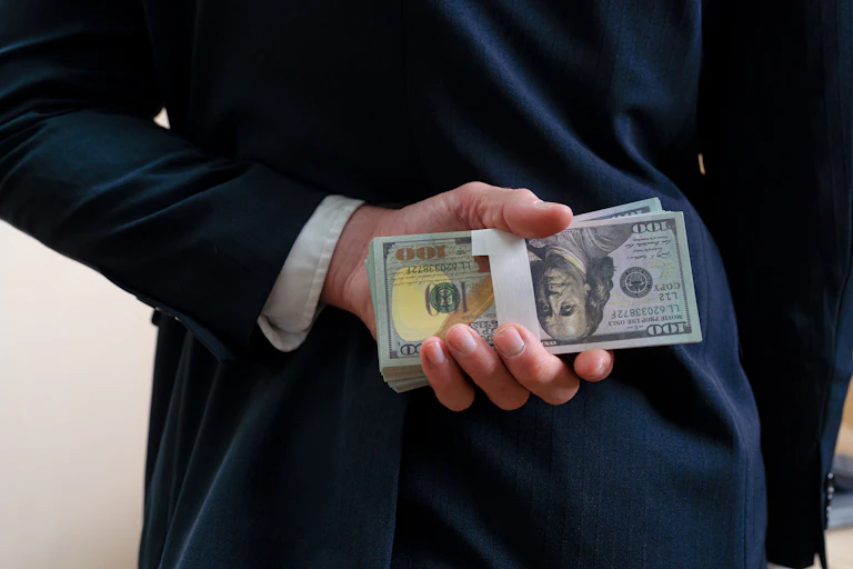 A businessman in a suit holds a stack of hundred-dollar bills behind his back.