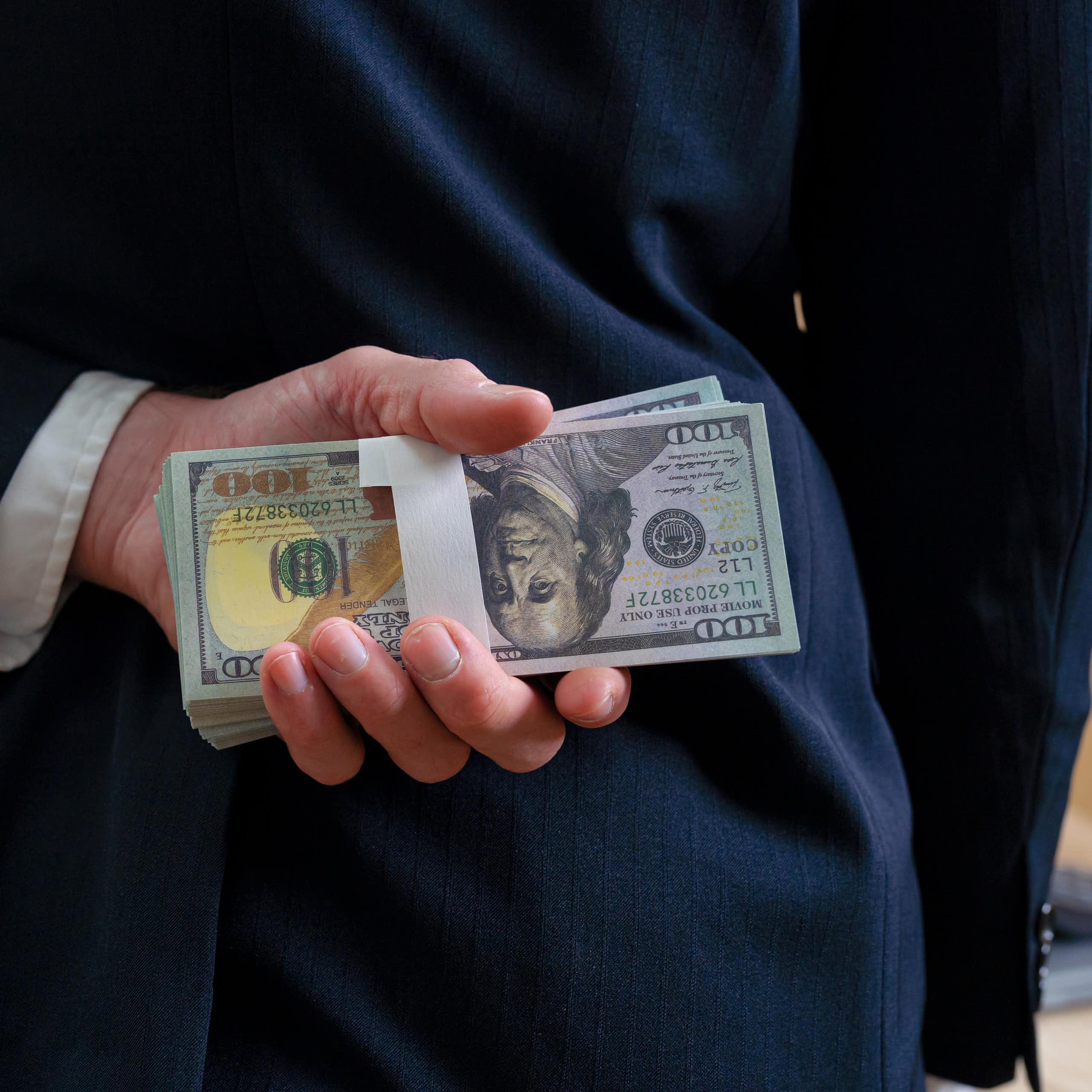 A businessman in a suit holds a stack of hundred-dollar bills behind his back.