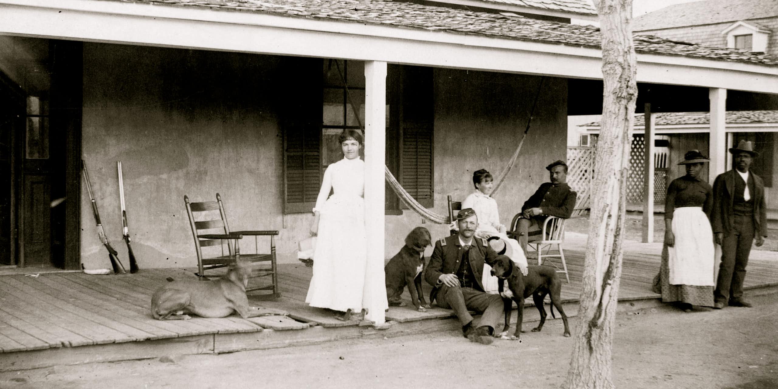A black and white photo shows a white woman standing against a pillar, with two soldiers and dogs and another woman seated nearby her. A Black man and woman stand off to the side. 