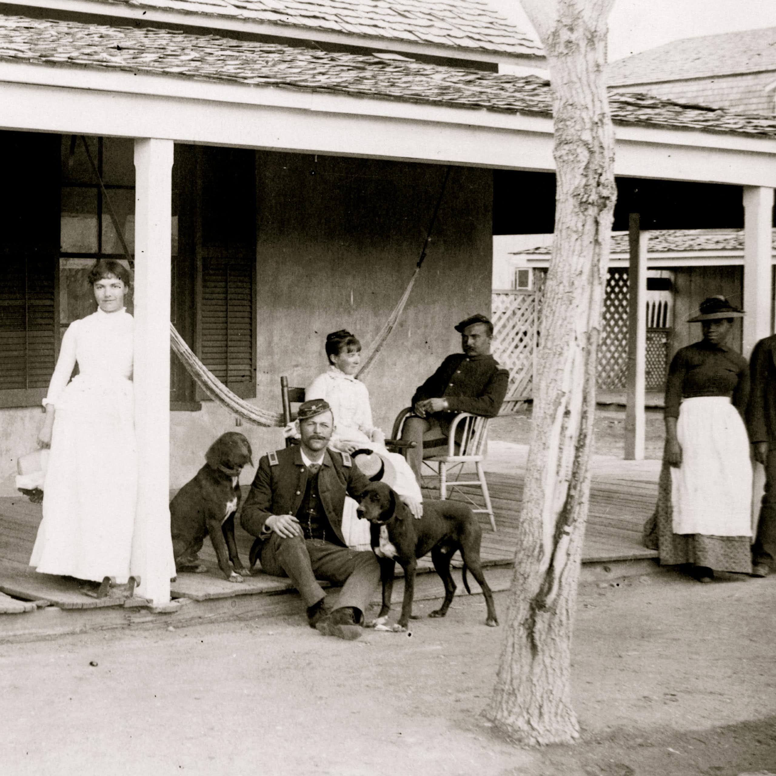A black and white photo shows a white woman standing against a pillar, with two soldiers and dogs and another woman seated nearby her. A Black man and woman stand off to the side. 