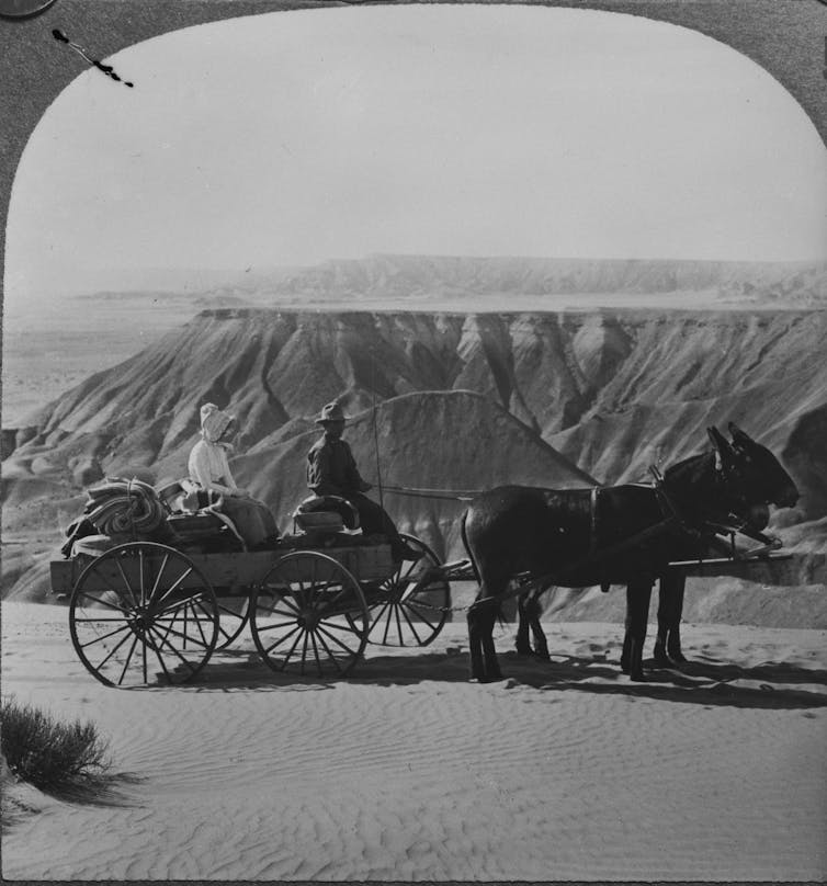 A black and white photo shows a man and a woman on a carriage pulled by two horses overlooking a vast desert landscape.