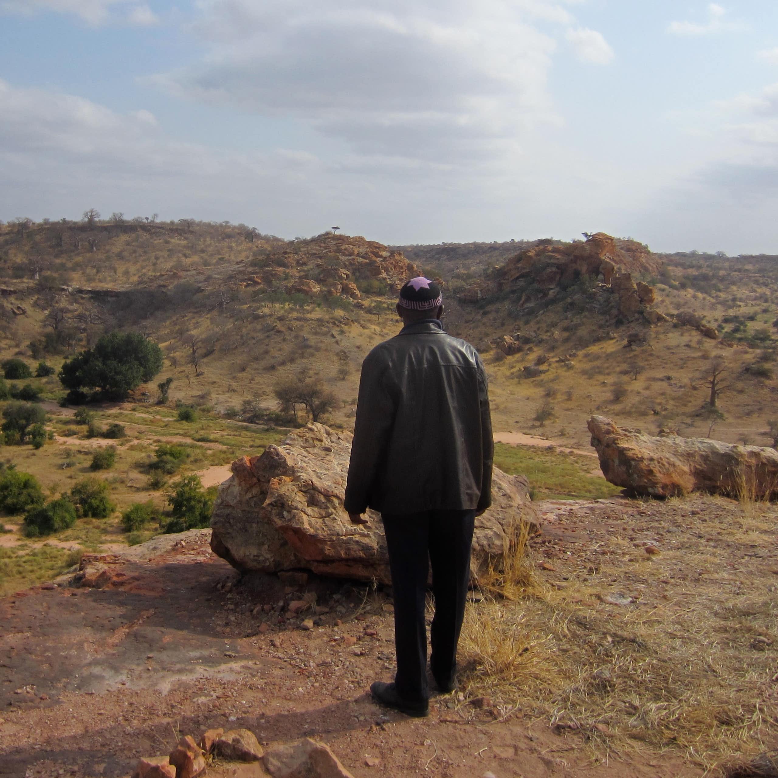 A man wearing black clothes and a yarmulke looks out across a mountainous landscape