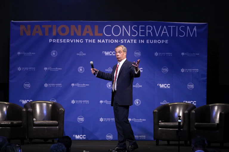 A man on stage with a microphone in his hand gestures as he looks to the audience. A National Conservatism banner is behind him.