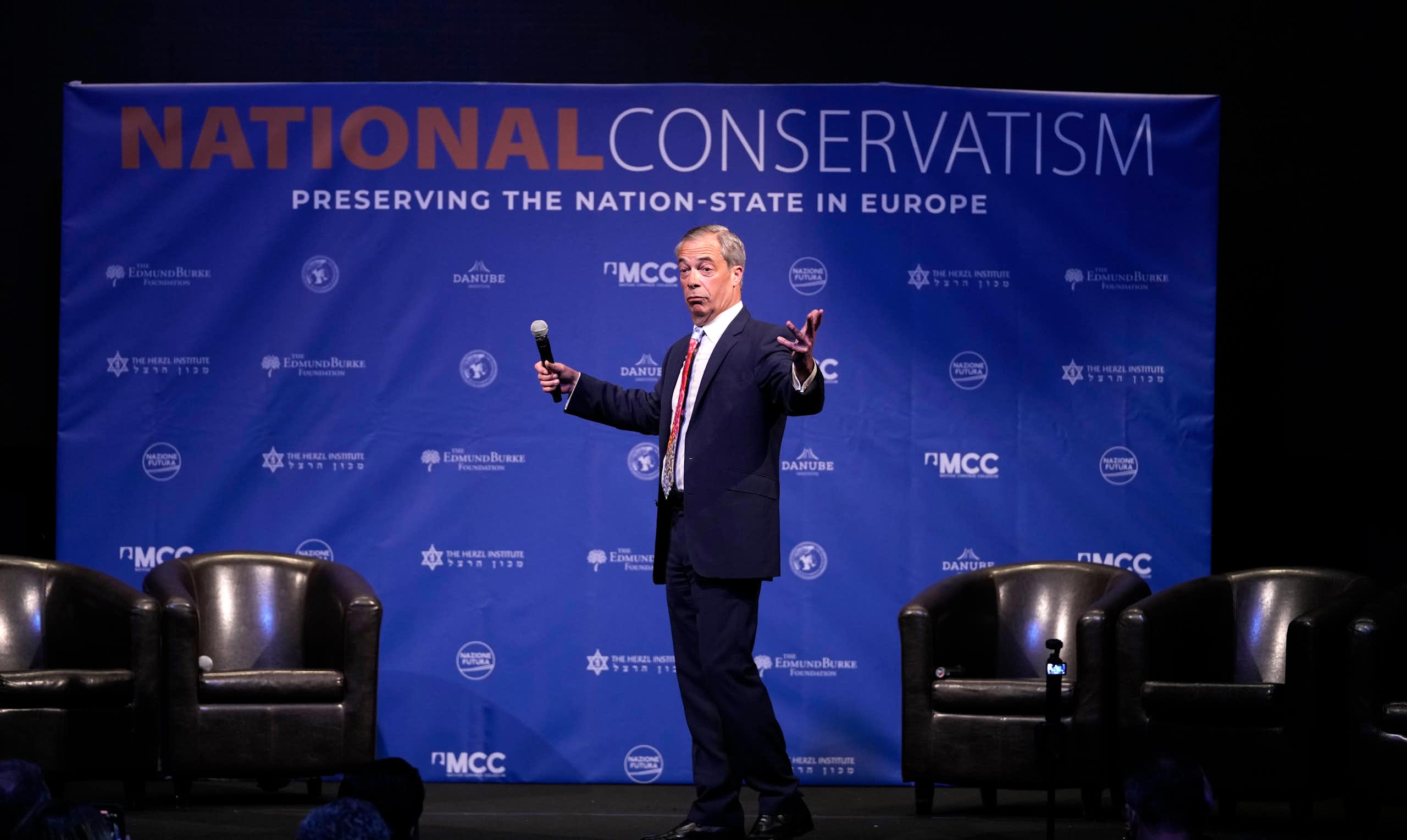 A man on stage with a microphone in his hand gestures as he looks to the audience. A National Conservatism banner is behind him.