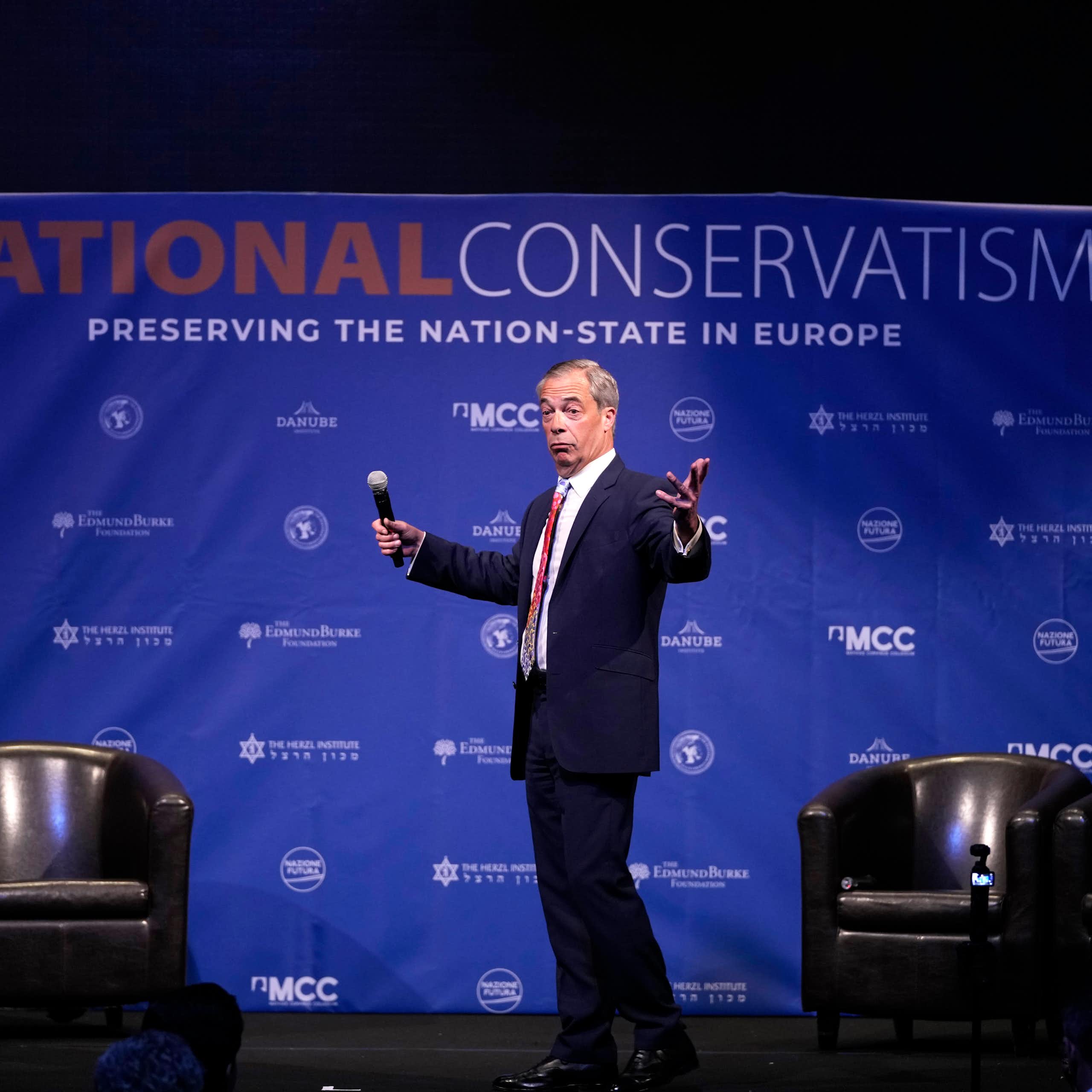 A man on stage with a microphone in his hand gestures as he looks to the audience. A National Conservatism banner is behind him.