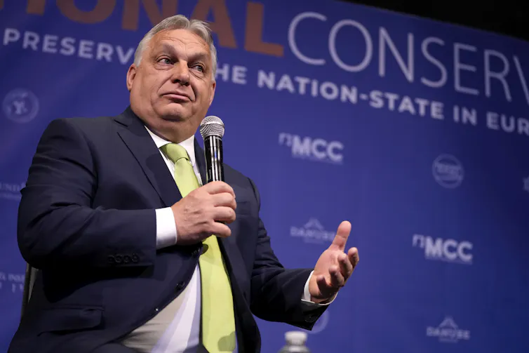 A rotund man in a yellow tie gestures while holding a microphone.