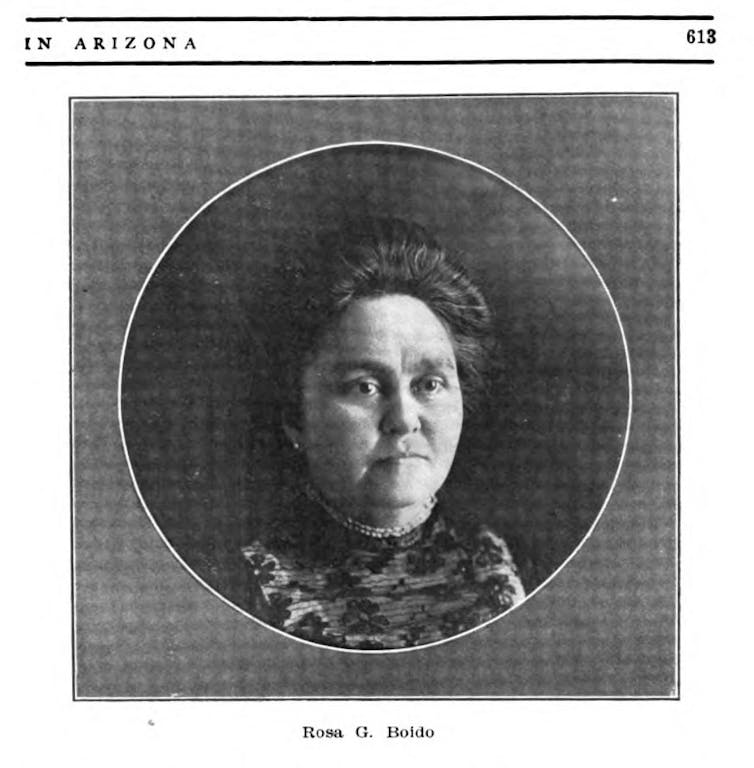 A black and white photo shows a woman seen from the head down to the chest. She has dark hair and wears formal clothing.