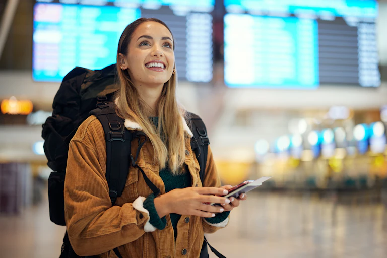 A young woman wearing a backpack and holding her passport smiles up at a screen in an airport