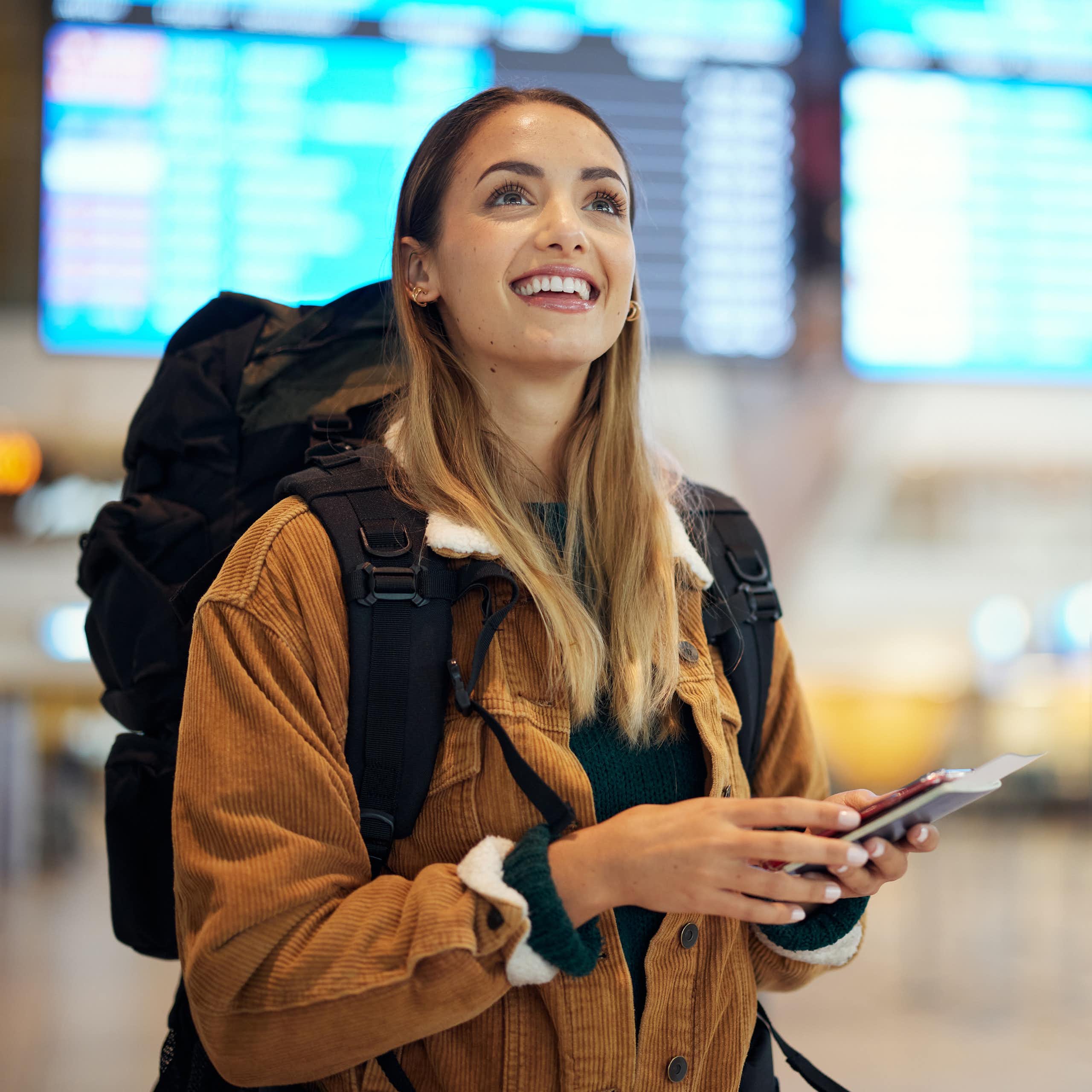 A young woman wearing a backpack and holding her passport smiles up at a screen in an airport