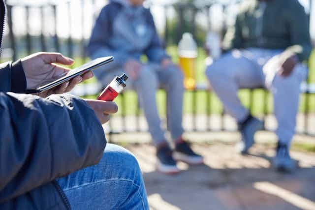 Primer plano de una mano sujetando un vapeador y la otra un teléfono móvil. 