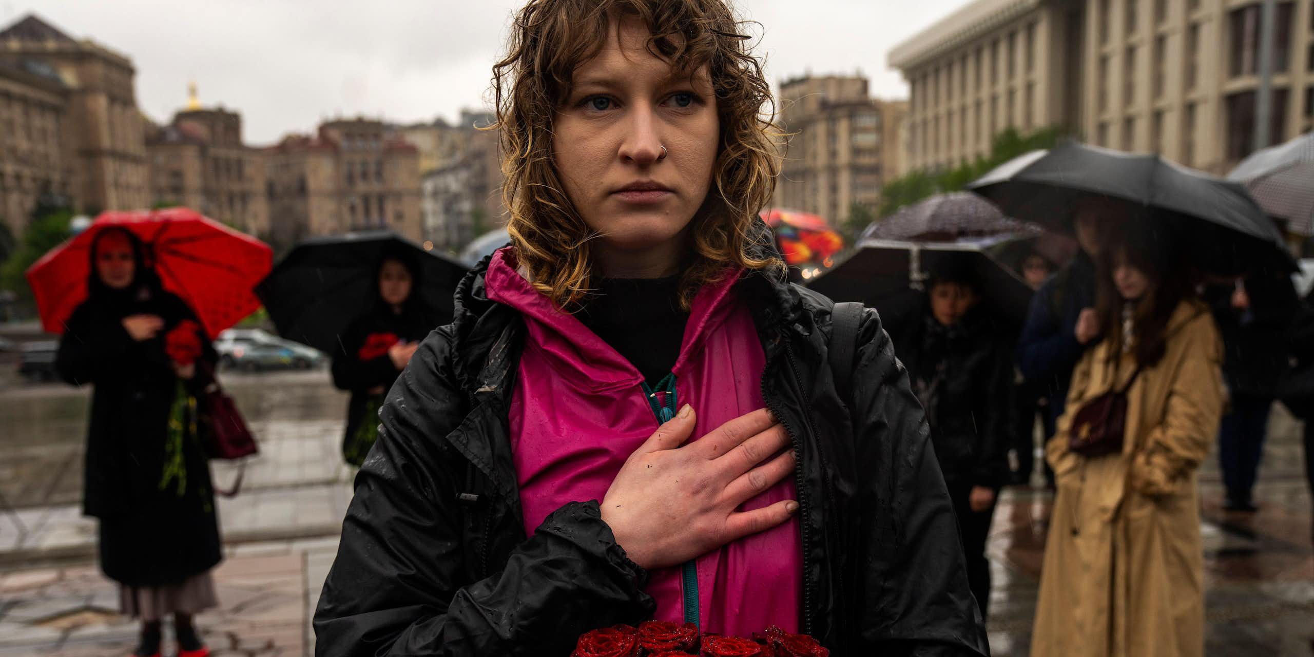 A yong woman stands in the rain at a funeral in KHarkiv, Ukraine, with her hand on her heart.