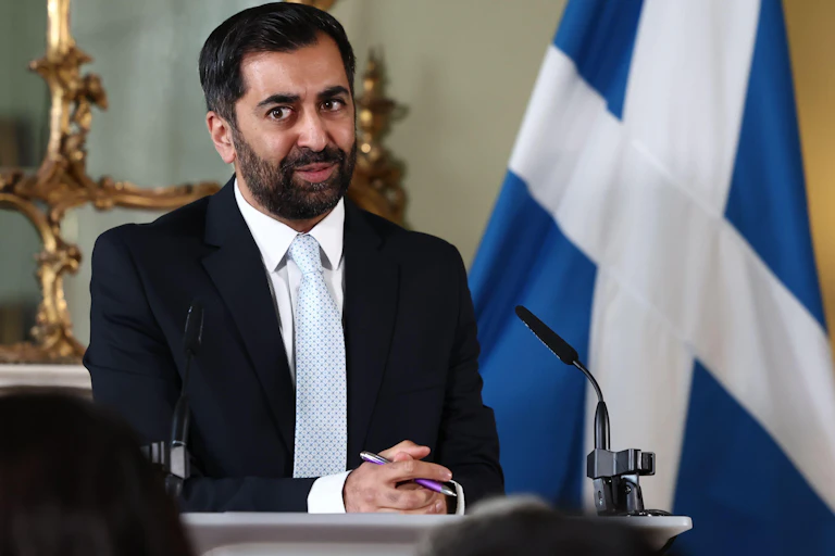 Humza Yousaf giving a speech in front of a Scottish flag.