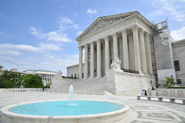 The Supreme Court building with white pillars is seen from an angle, with a small pool and fountain in front of it on a blue-sky day.