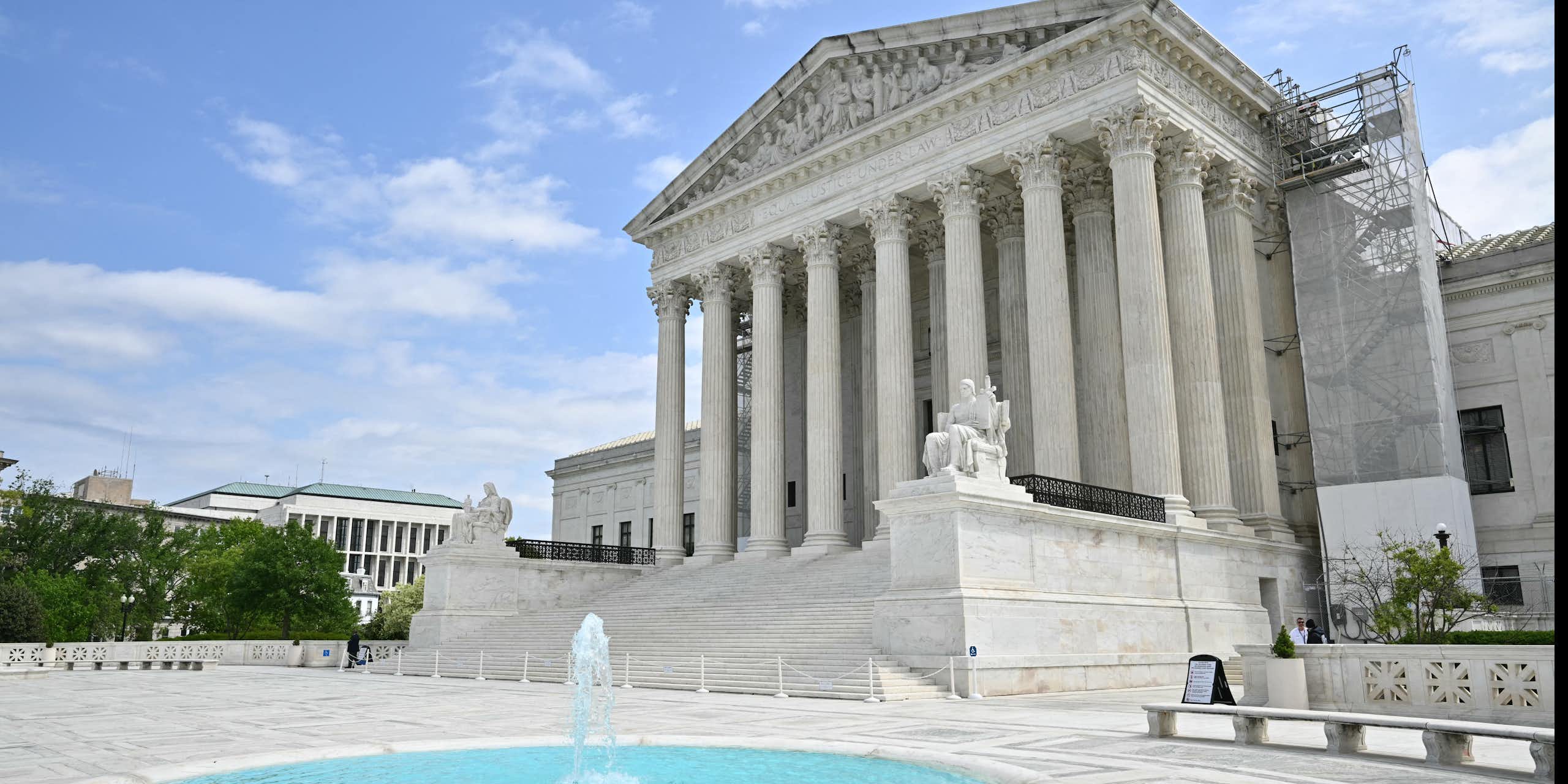 The Supreme Court building with white pillars is seen from an angle, with a small pool and fountain in front of it on a blue-sky day.