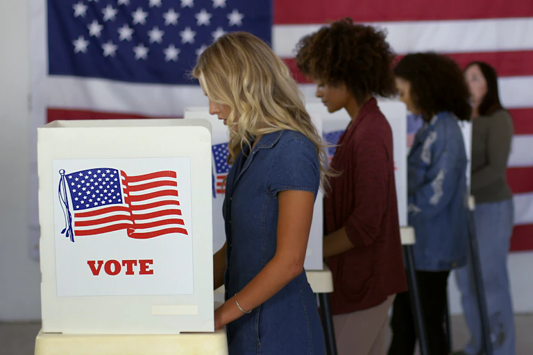 Four young women of different ethnicities, filling in ballots and casting votes in booths at polling station, with a US flag on the wall behind them.
