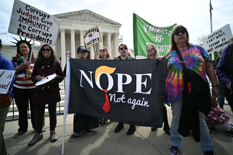 A group of people stand in front of the Supreme Court holding signs that read, 