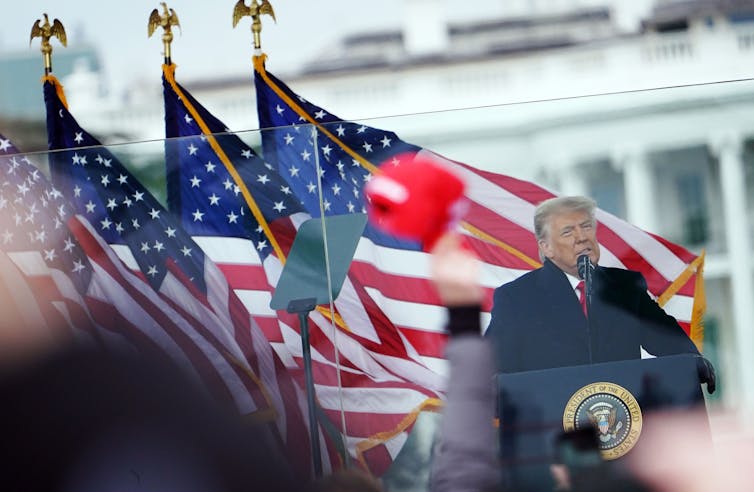 A white man in a dark jacket stands at a podium in front of a white building and a row of American flags.