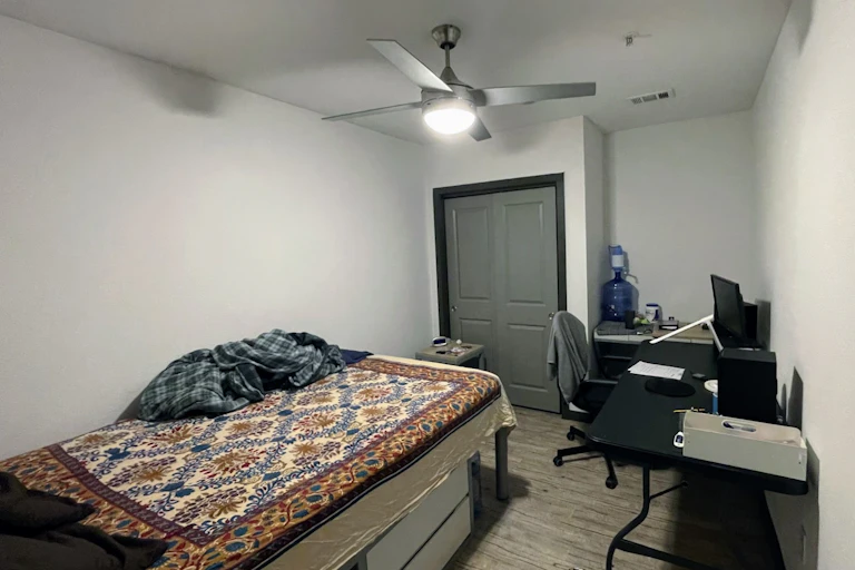 A windowless room features a single twin bed, a desk and a chair.