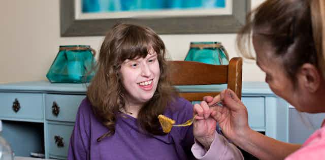 Disabled young woman is assisted to use her fork to eat dinner.