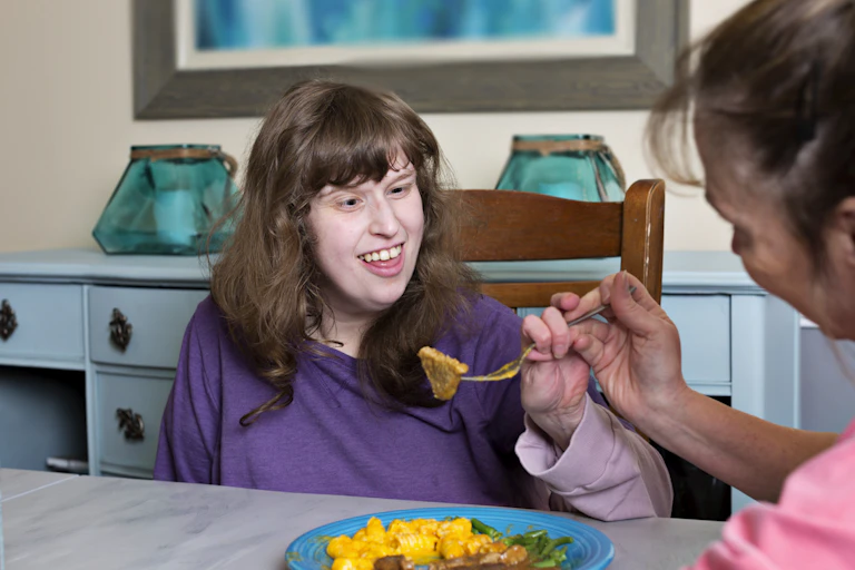 Disabled young woman is assisted to use her fork to eat dinner.