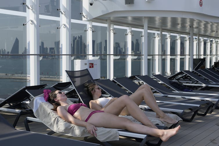 Two women in bathing suits lie on reclined lounge chairs on the deck of a ship