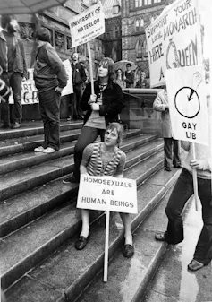 A black and white photo of people protesting on building steps. One placard reads: homosexuals are human beings.