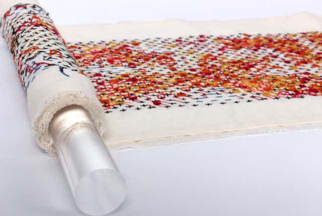 an embroidered scroll in an abstract pattern in black, red, orange and yellow wound a clear perspex rod.