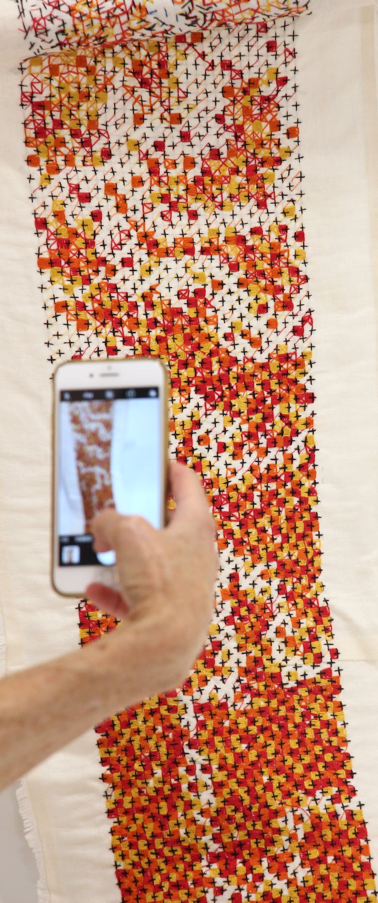 A hand holds a phone taking a picture of a long abstract embroidery in black, red, orange and yellow.