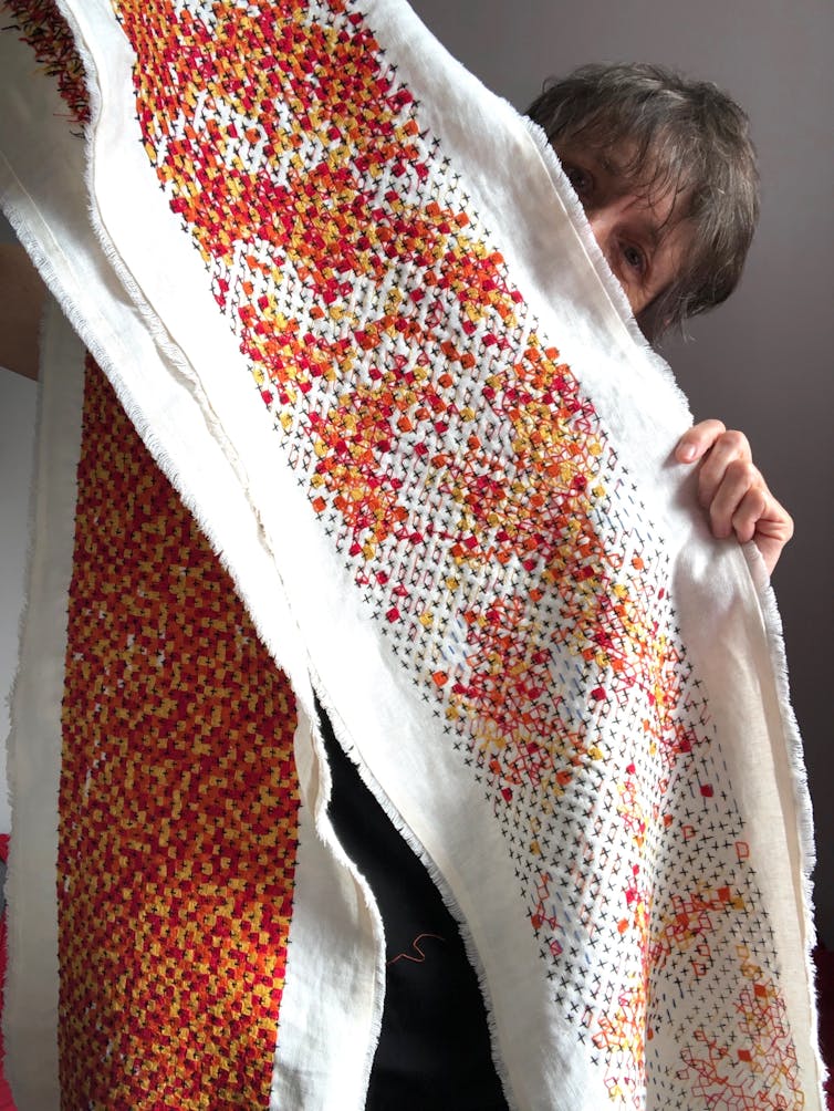 A middle-aged white woman peeks out from behind a very long abstract embroidery in black, red, orange and yellow.