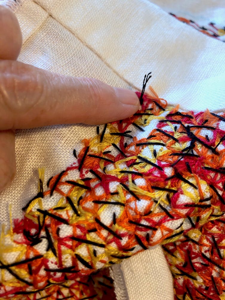A finger points to a knot on the back of a messy abstract embroidery done in black, red, orange and yellow