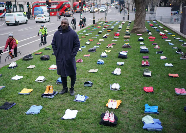 A man stands on a green surrounded by piles of neatly folded clothes.