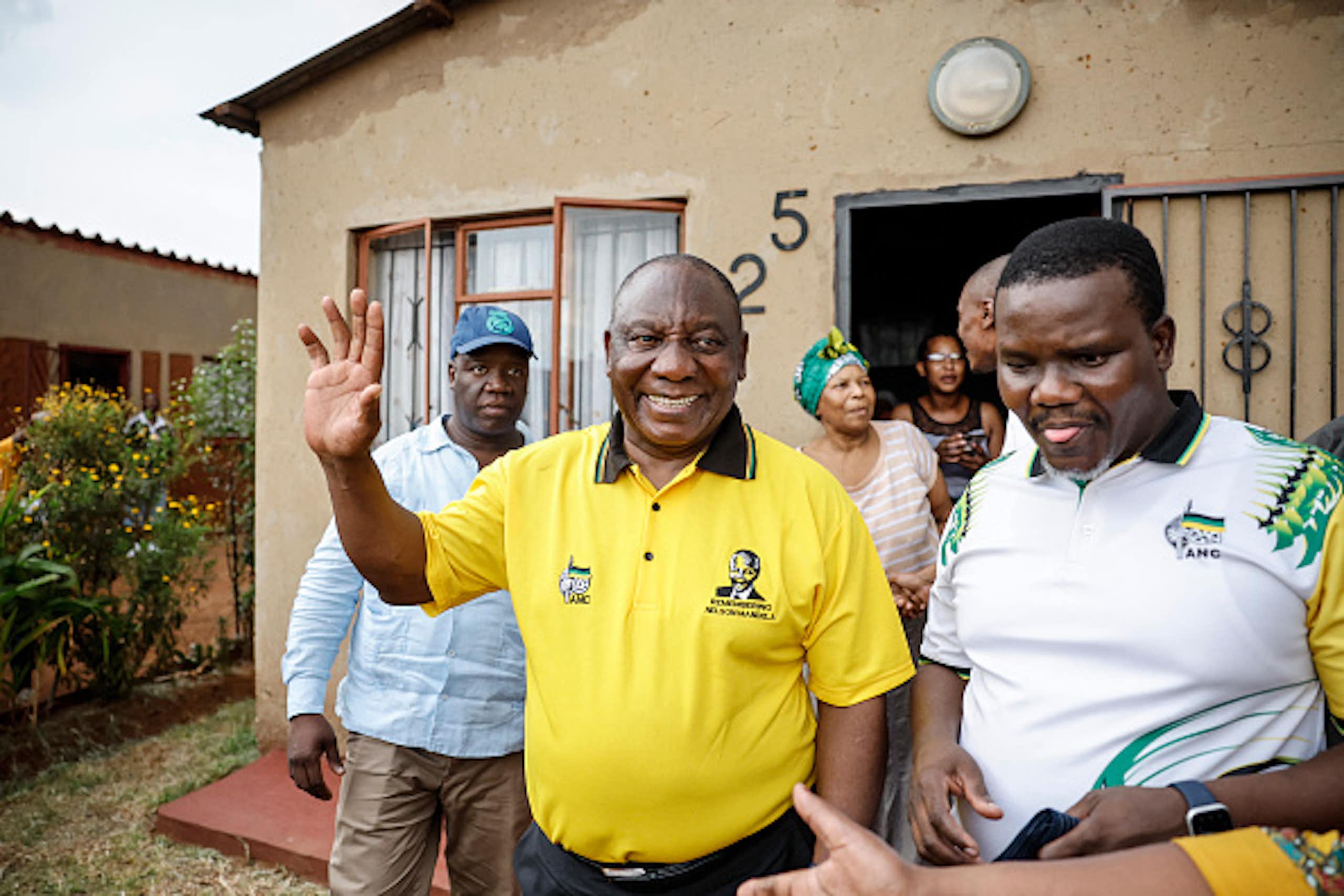 A man in a yellow short smiles and raises a hand in greeting outside a house, with other people standing next to and behind him