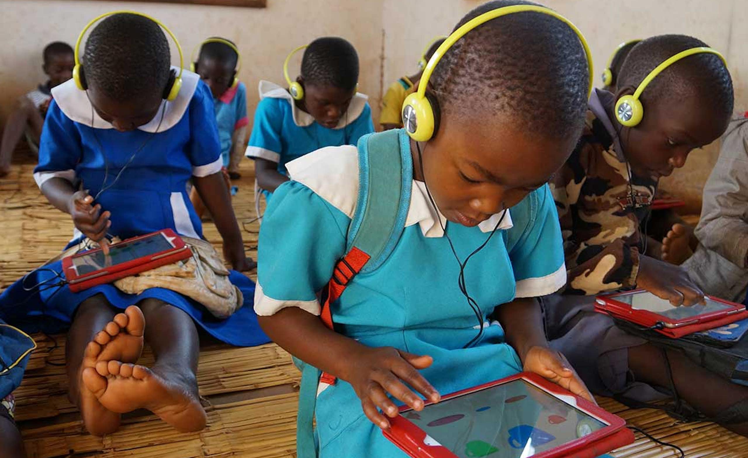 A group of children, most in blue school uniforms and yellow headphones, work on red computer tablets